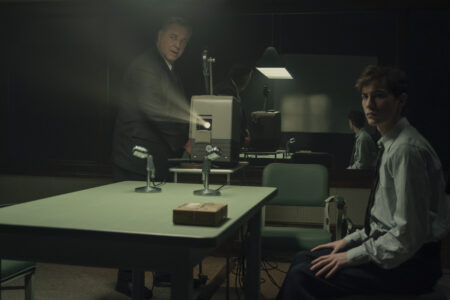 "Two men in formal attire are in a dimly lit interrogation room with a green table, microphones, and a film projector. One man stands by the projector while the other sits, suggesting a tense interview or investigation setting." Unabomber. (L to R) Russell Crowe as Henry Murray and Jacob Tremblay as Ted Kaczynski in Unabomber.