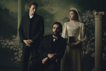 "Three people in Victorian-era clothing pose in a formal portrait setting with floral and classical column background; one man seated, one man standing, and a woman in a white dress holding a book." East of Eden. (L to R) Mike Faist as Charles Trask, Christopher Abbott as Adam Trask, Florence Pugh as Cathy Ames in episode 103 of East of Eden