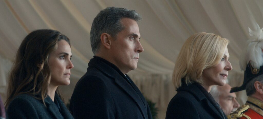 "Three serious-looking adults in formal dark clothing stand side by side in an elegant, tented environment. They are watching something intently, possibly at an official event or ceremony. A uniformed figure is partially visible." The Diplomat. (L to R) Keri Russell as Kate Wyler, Rufus Sewell as Hal Wyler, Allison Janney as Grace Penn in episode 401 of The Diplomat.